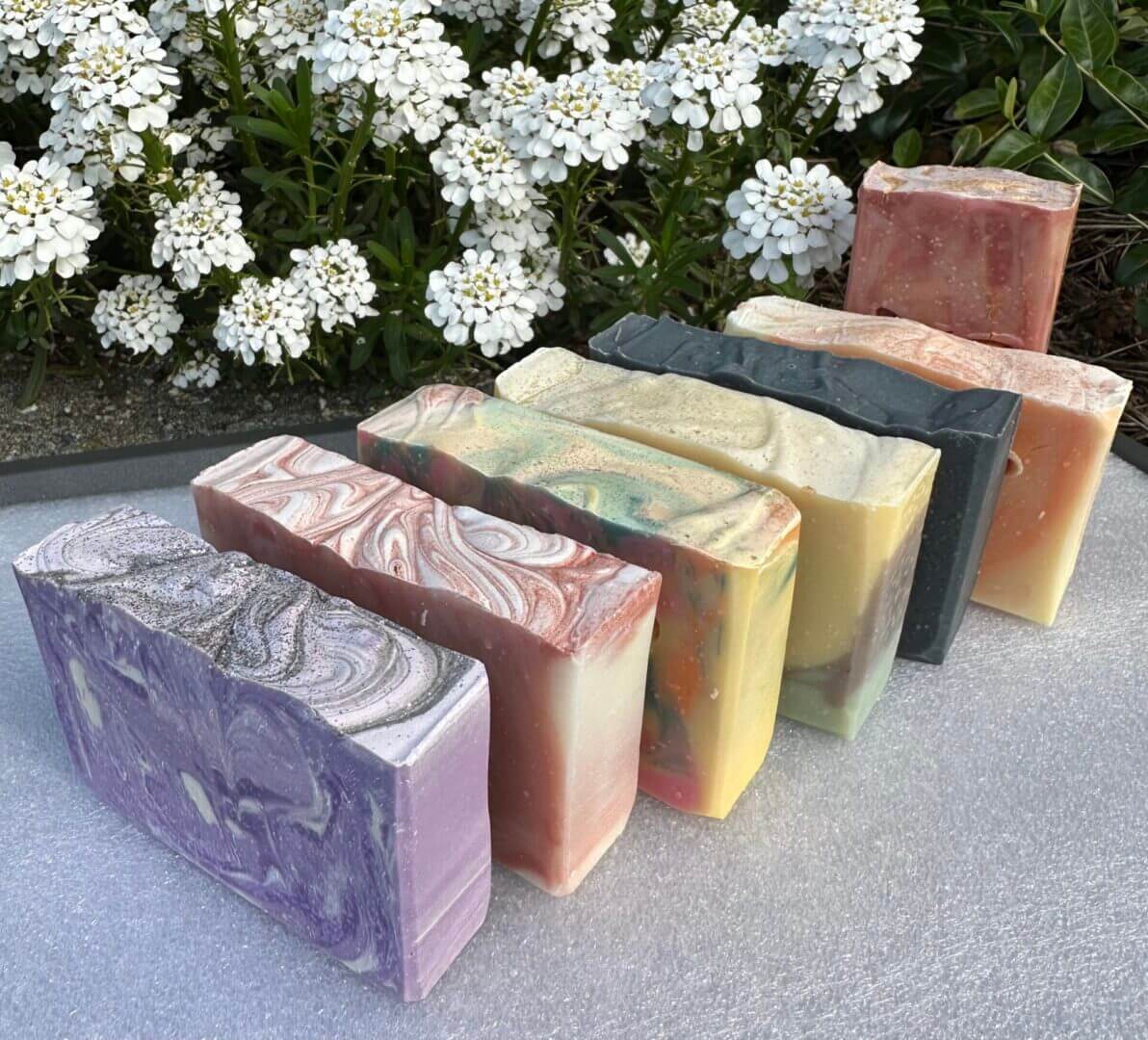A row of colorful handmade soaps placed outdoors near white flowers.
